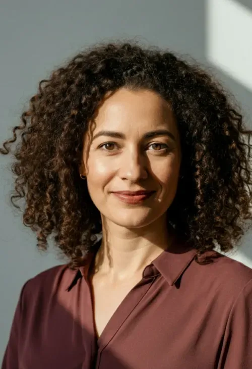 Smiling woman with curly hair wearing a maroon button-up shirt against a neutral background.