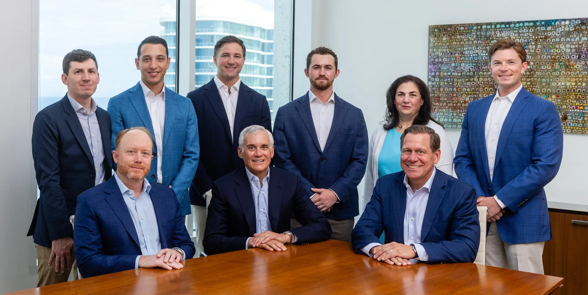 Group of nine business professionals posing in a modern office around a wooden conference table.
