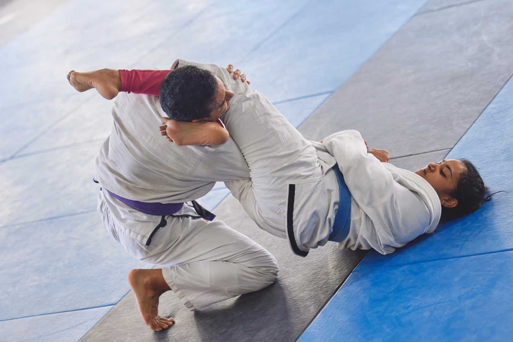 Two martial artists practicing Brazilian jiu-jitsu on mats, one applying a guard technique.