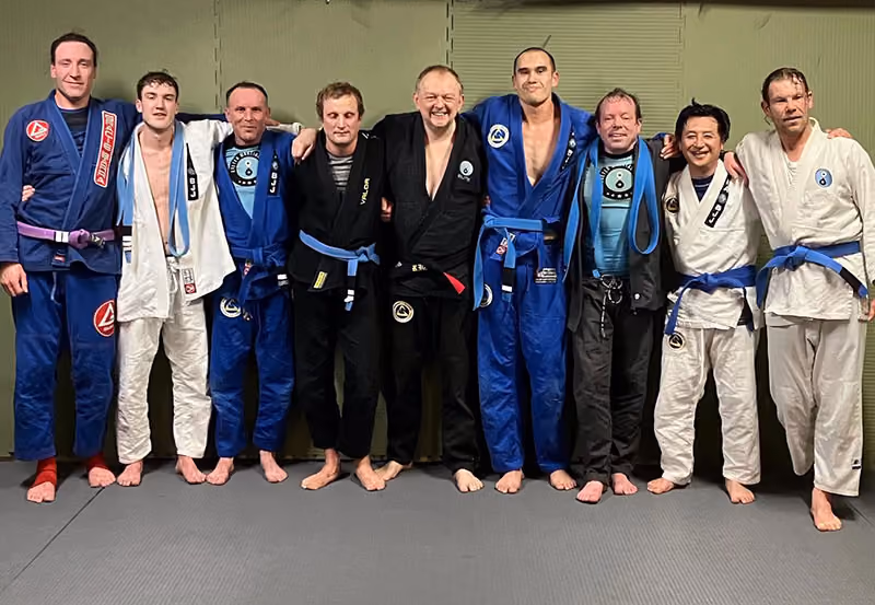 Group of nine men standing on mats wearing varied Brazilian jiu-jitsu gis with different belt colors.