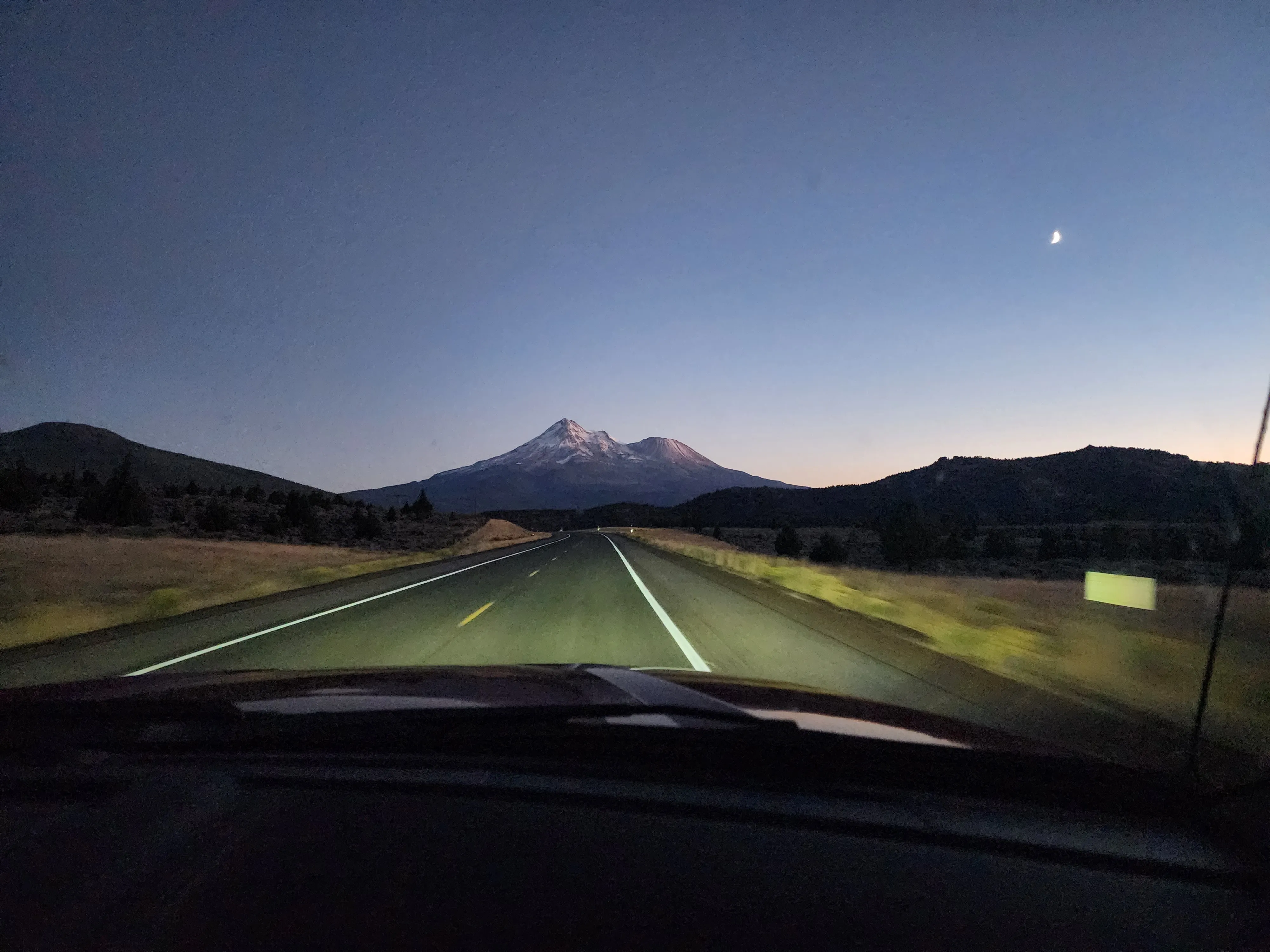 Mountain road leading toward snow-covered Mount Shasta beneath a deep blue twilight sky, where a distant glow beyond the summit evokes growth, momentum, and acceleration.