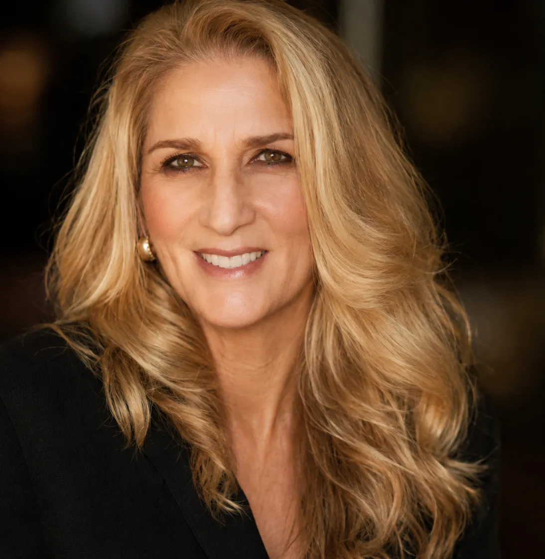 Portrait of Rebecca Camille smiling warmly, wearing a black blazer with softly curled blonde hair against a dark background.