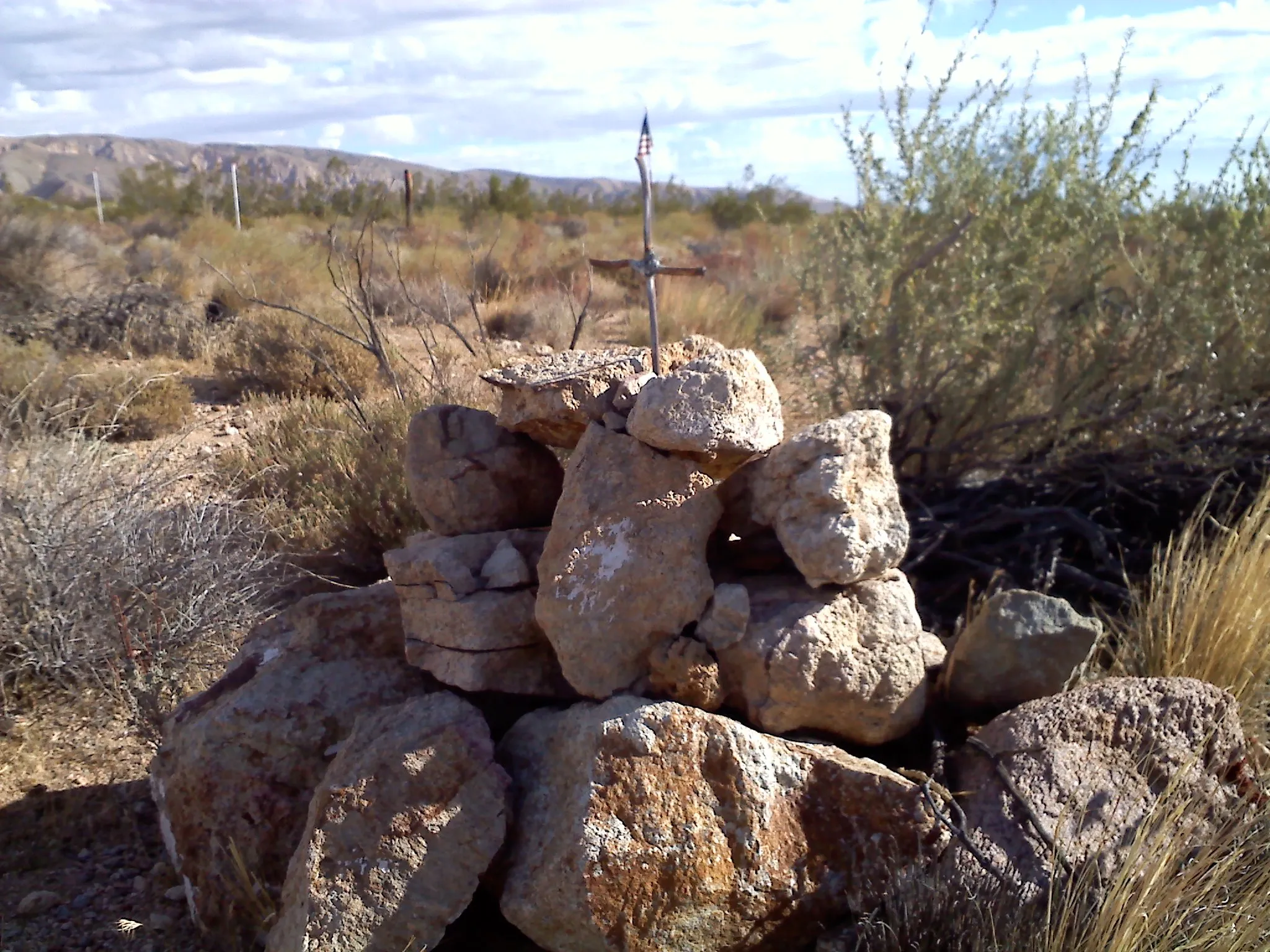 "Rock cairn with a cross marking a desert location, surrounded by Mojave shrubs with distant mountains beneath a cloudy sky."