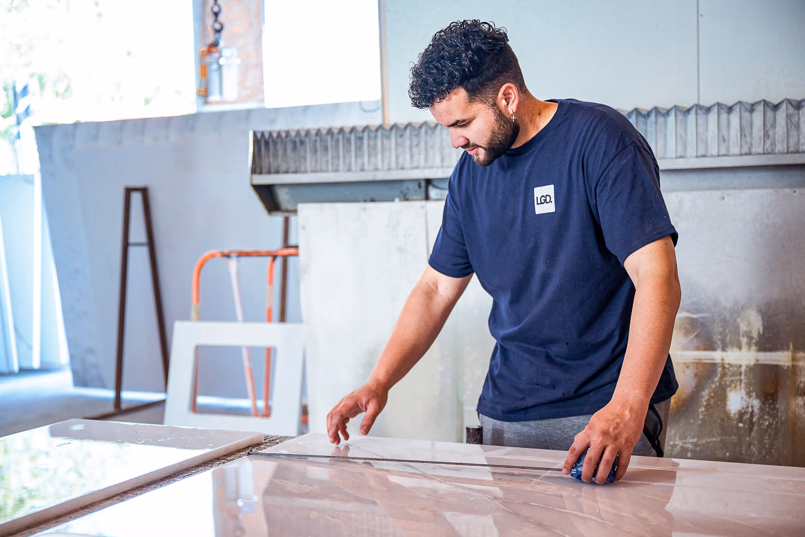 Man wearing navy LGD shirt inspecting or measuring a large polished stone slab indoors.