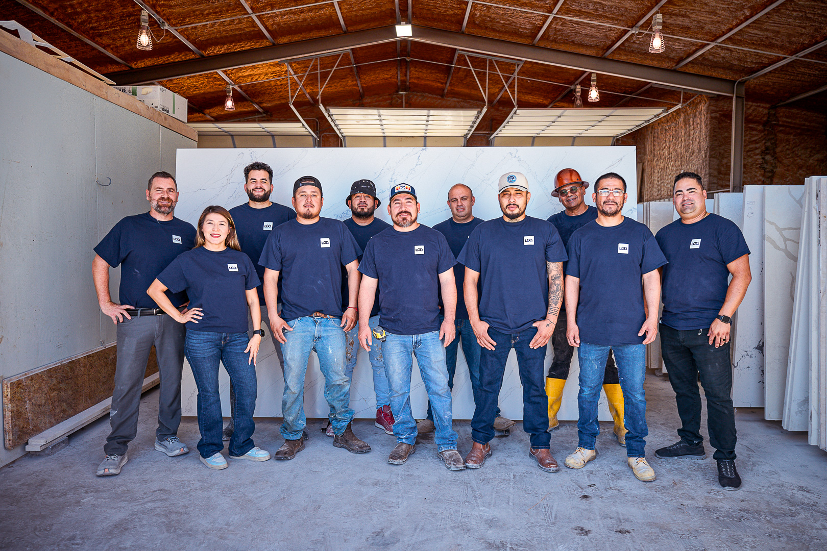 Group of twelve people wearing matching navy shirts standing in a warehouse with marble slabs behind them.