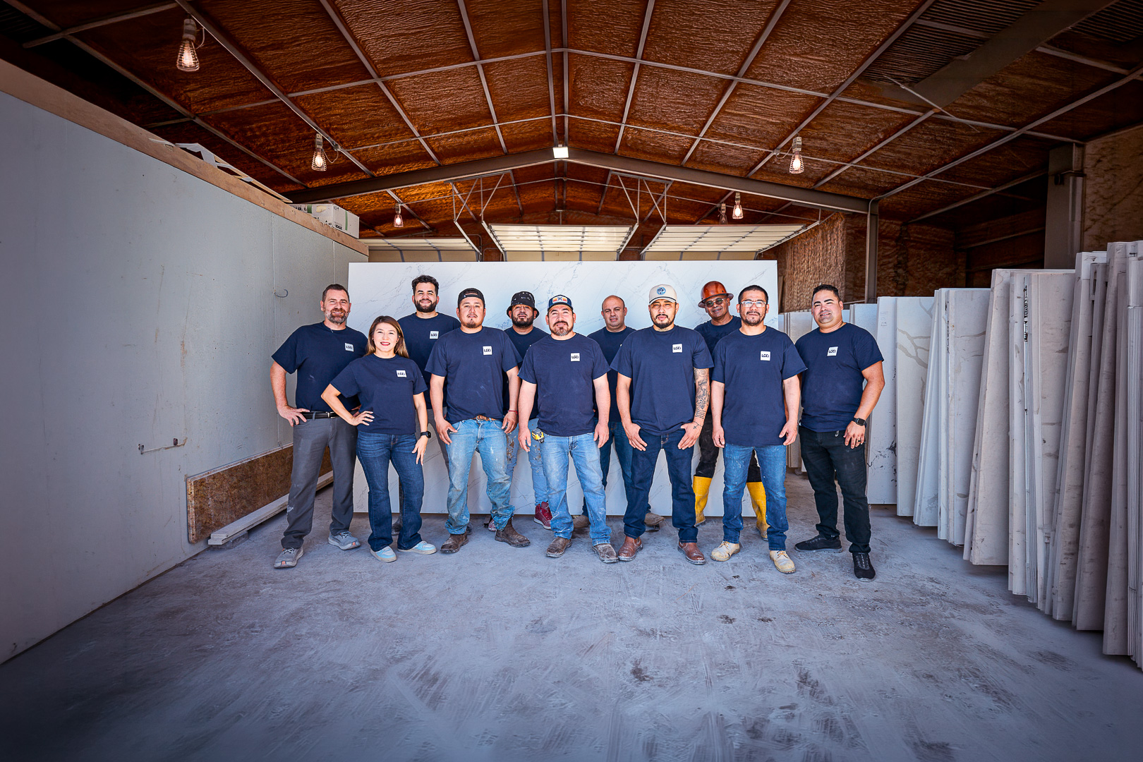 Group of 11 workers in navy shirts standing in a warehouse with marble slabs stacked around.