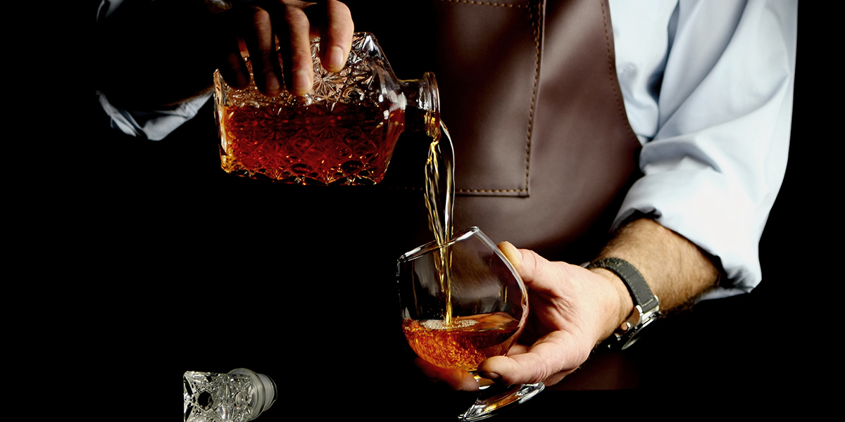 Person wearing brown apron pouring amber liquor from a cut-glass decanter into a tilted glass.