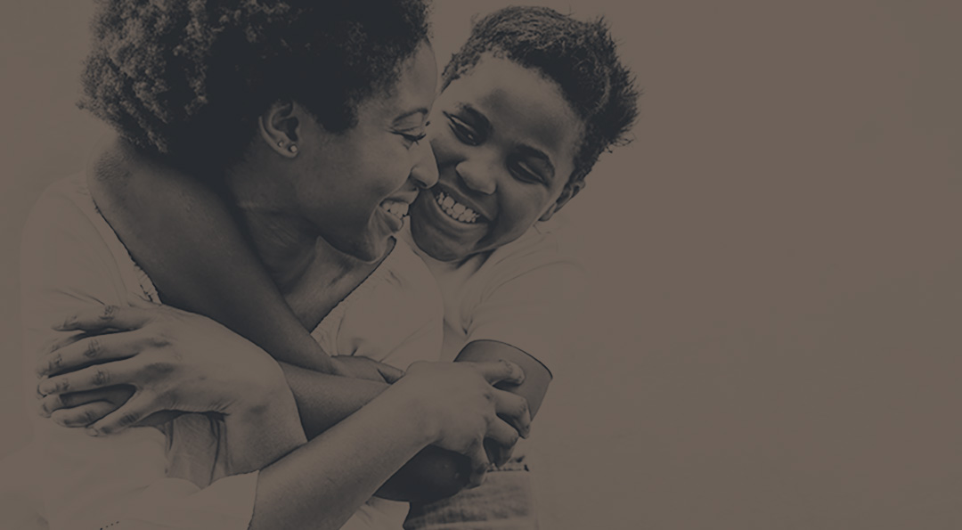 Smiling child hugging a joyful woman with short curly hair against a plain background.