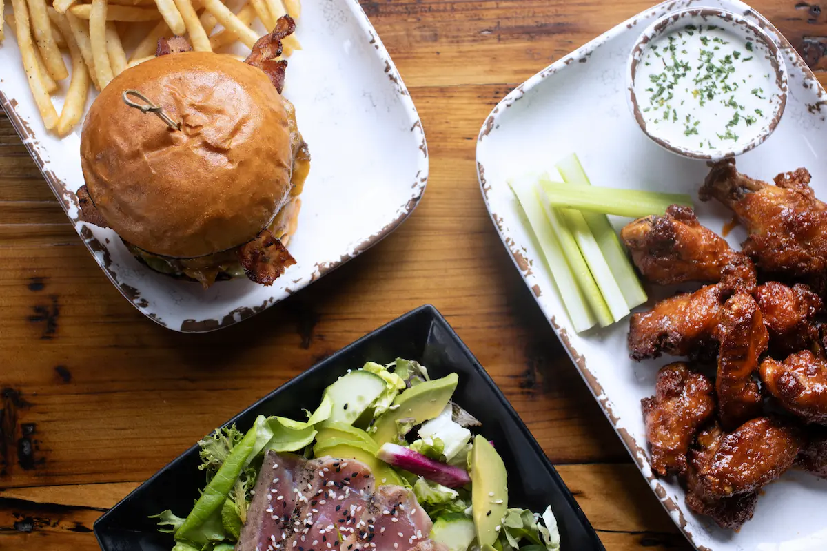 Top view of a meal with a cheeseburger and fries, a plate of buffalo chicken wings with celery and ranch dip, and a salad with seared tuna and avocado slices on a wooden table.