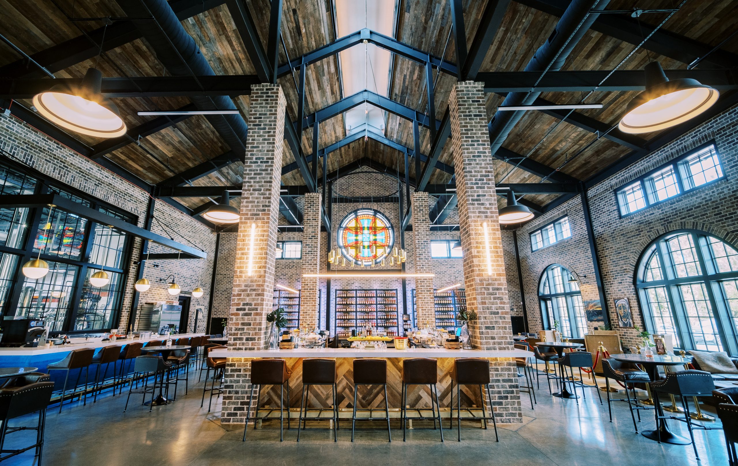 Spacious modern bar with wooden ceiling, brick pillars, large stained-glass window, and seating at the bar and tables.