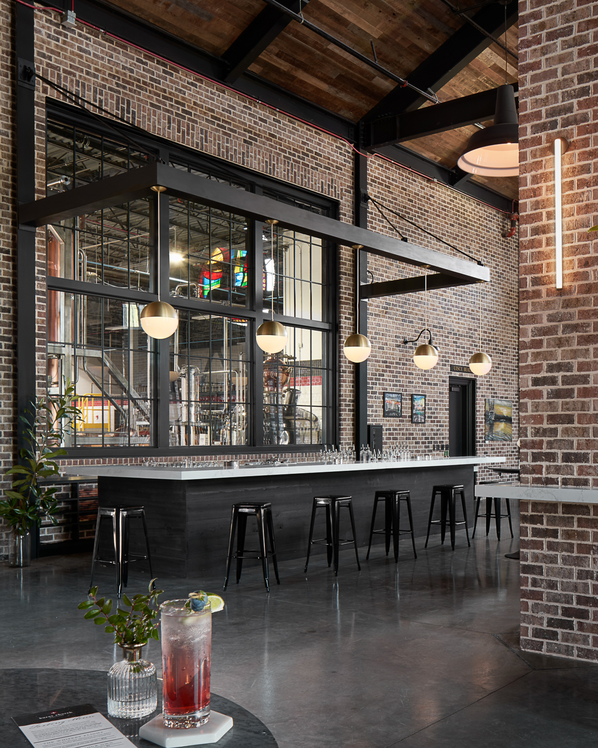 Modern bar interior with long white countertop, black stools, large industrial windows, and hanging globe pendant lights.