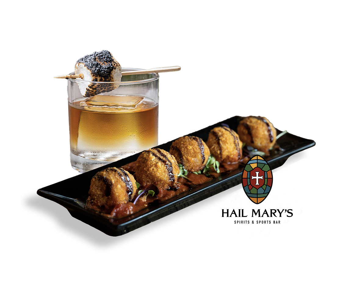 Glass of whiskey with ice and a toasted marshmallow on a skewer, next to a black tray of six fried appetizers drizzled with sauce on microgreens.