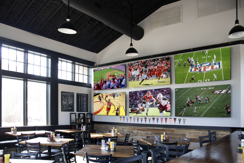 Interior of a sports-themed restaurant with multiple TV screens displaying baseball, football, and basketball games, wooden tables, and black chairs.