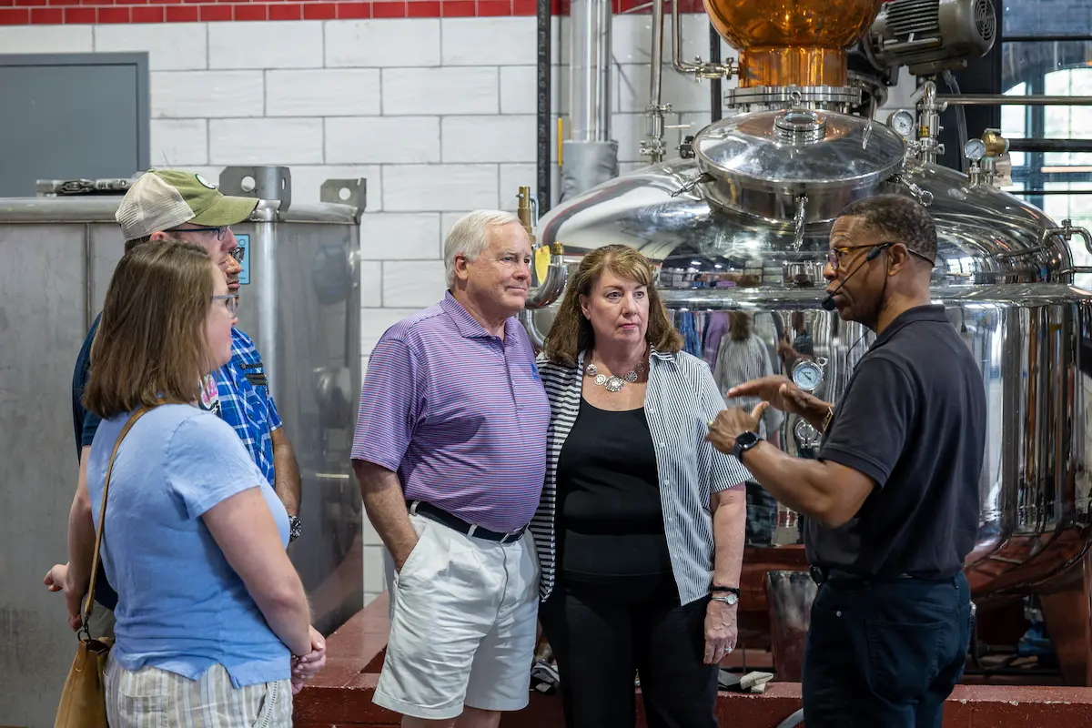 Group of adults listening to a guide speaking in front of large stainless steel brewing equipment inside a brewery.