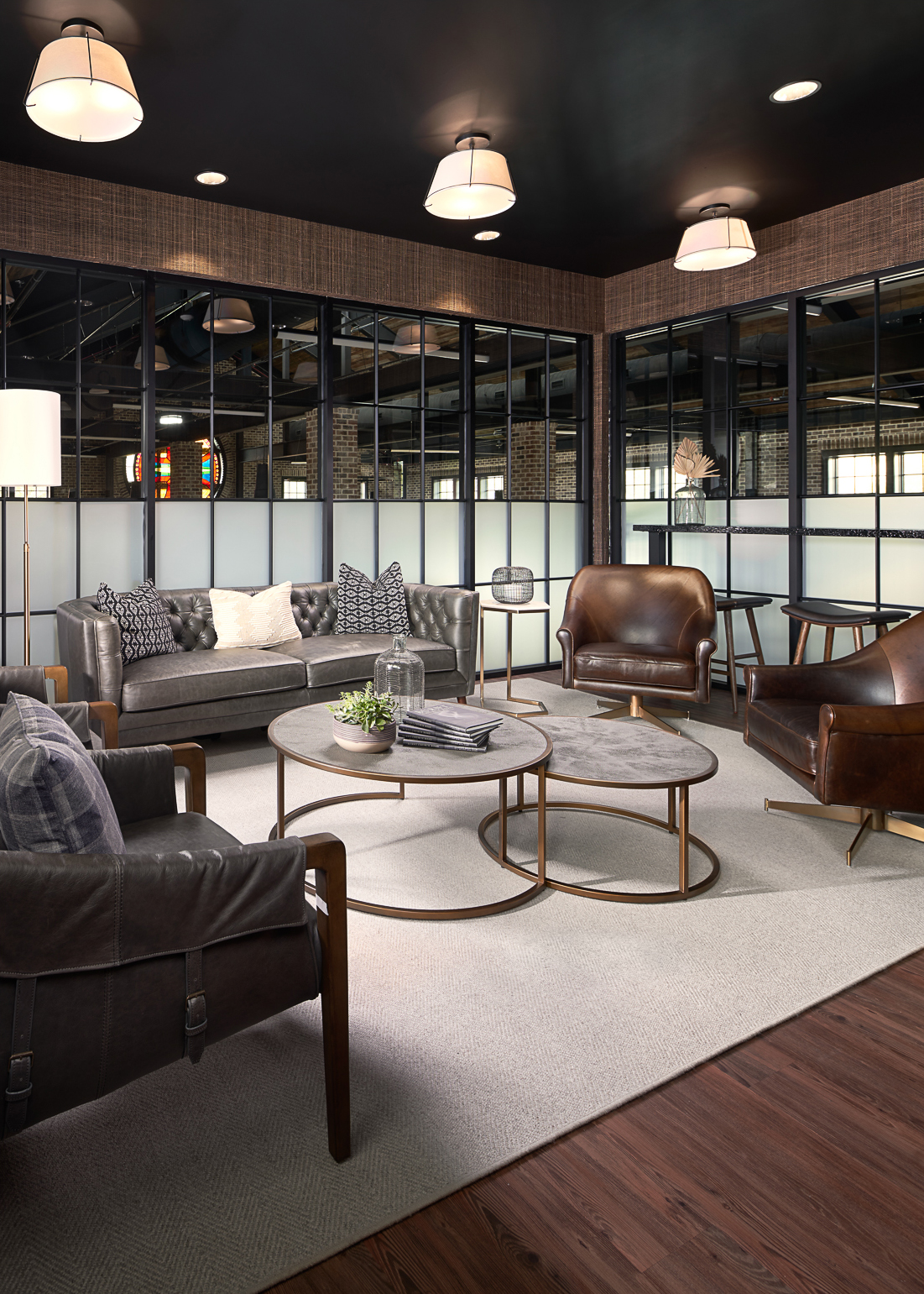 Modern lounge area with gray leather sofa, brown leather armchairs, two round coffee tables, and large windows with black frames.