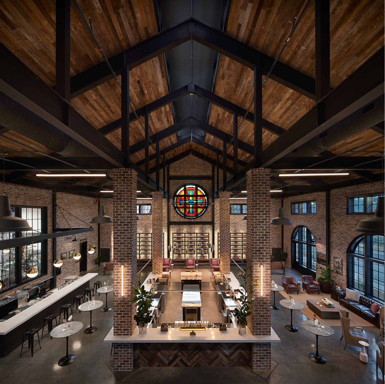 Spacious modern bar with high wooden ceiling, exposed brick walls, a large stained glass window, multiple seating areas, and marble-topped tables.