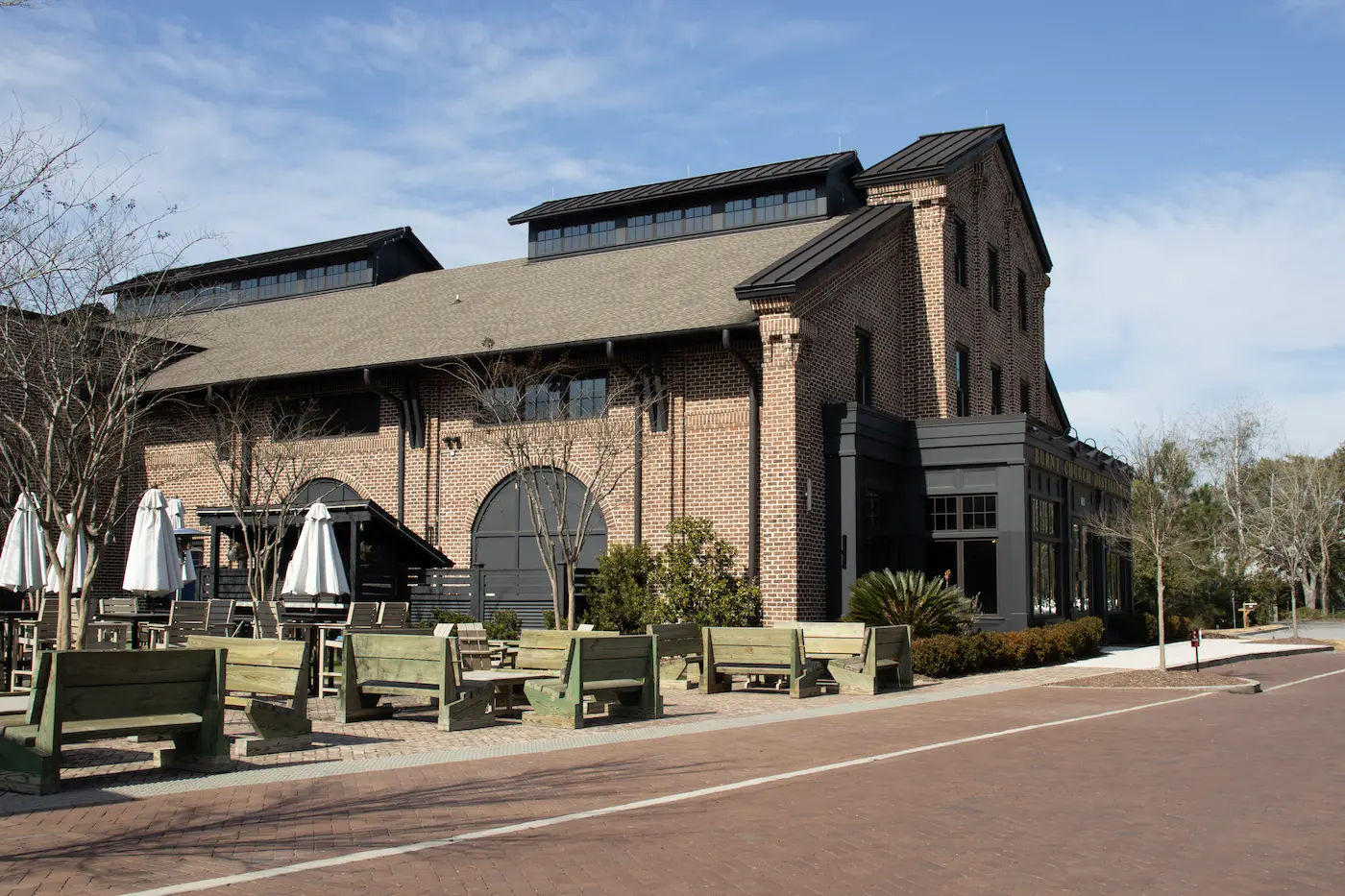 Large brick building with outdoor seating area featuring wooden benches and closed white umbrellas under a partly cloudy sky.