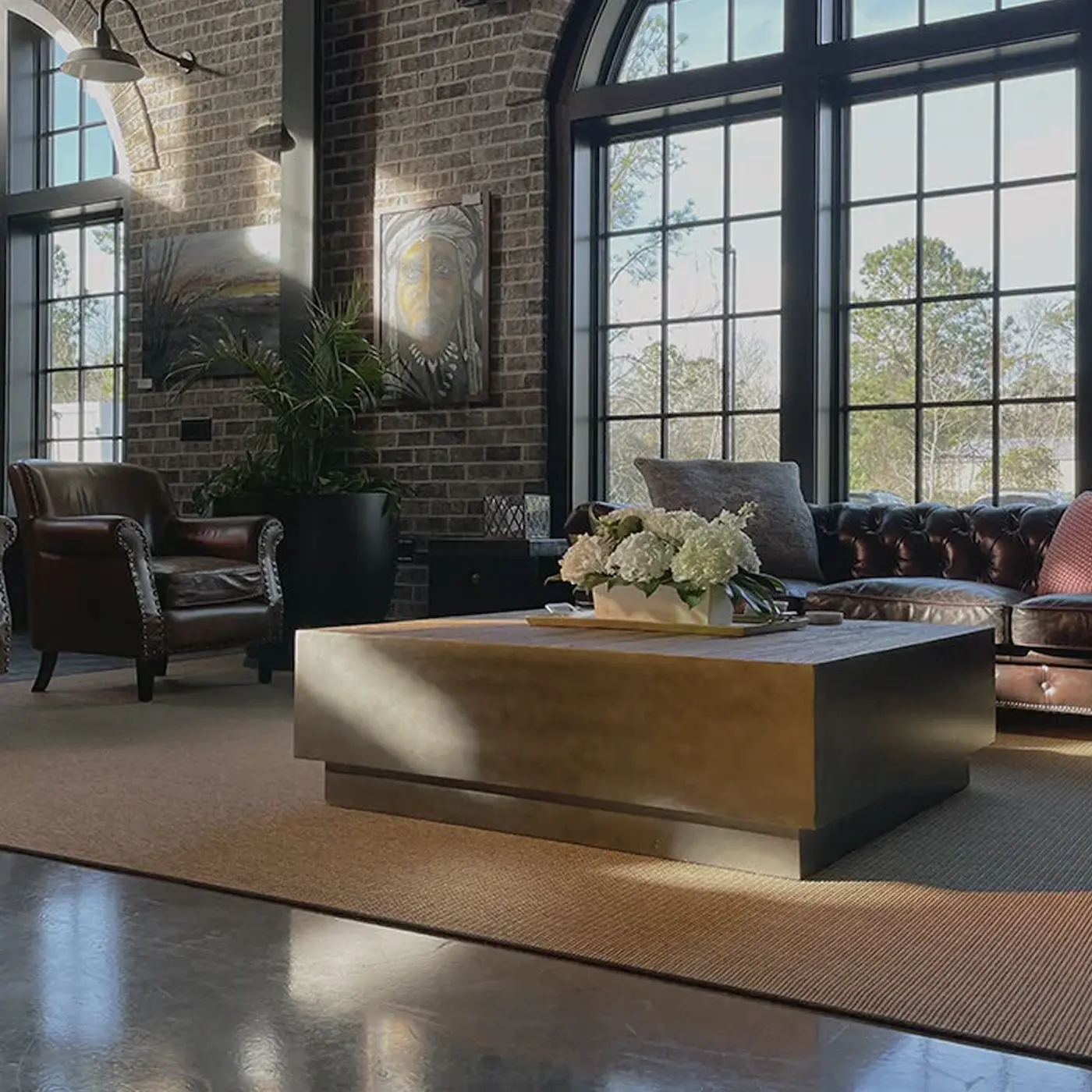 Sunlit living room with brown leather armchairs and sofa, large windows, brick wall, and a coffee table with white flowers.