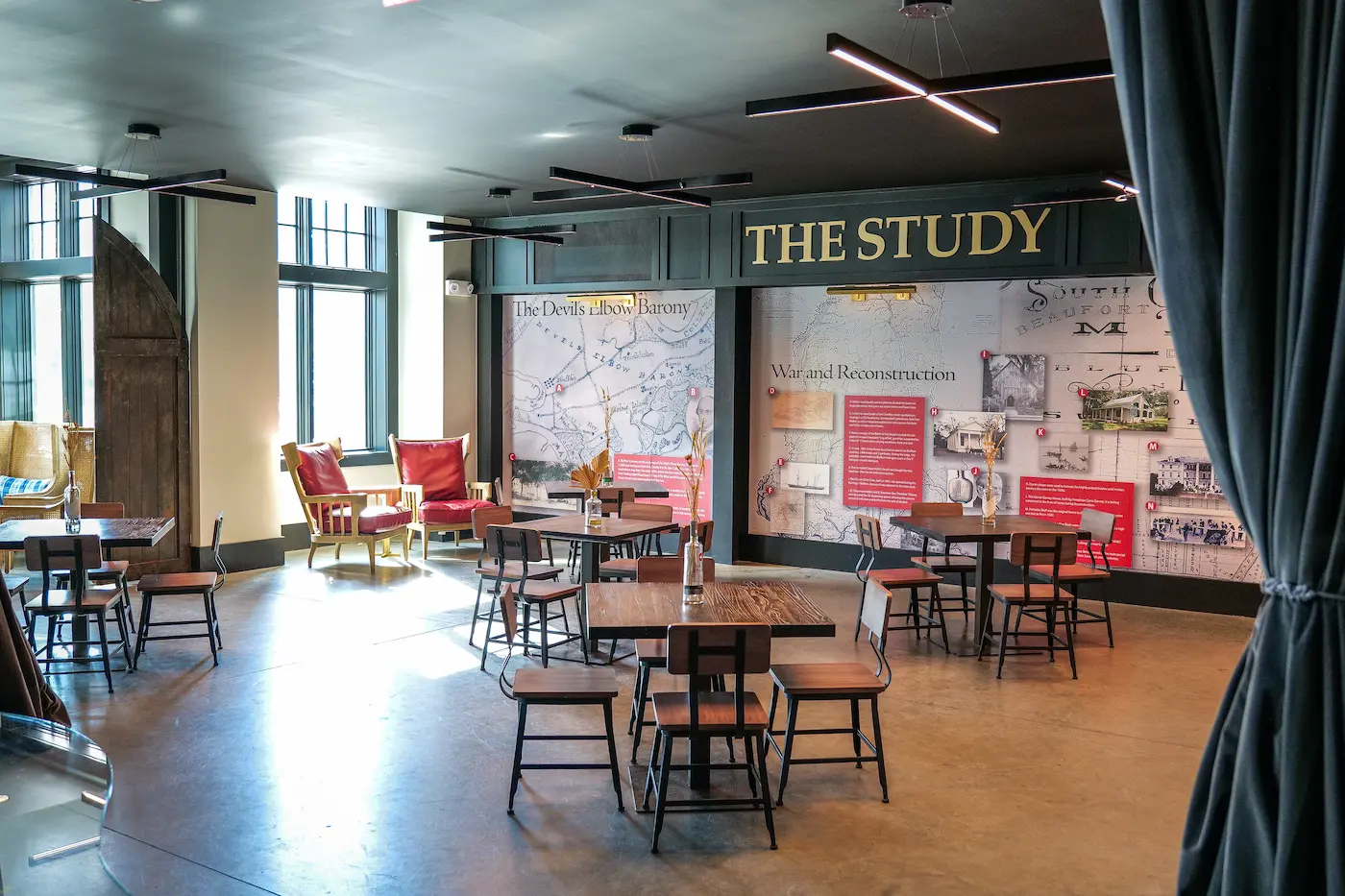 Light-filled study room with wooden tables and chairs, two red armchairs near windows, and historical wall displays.