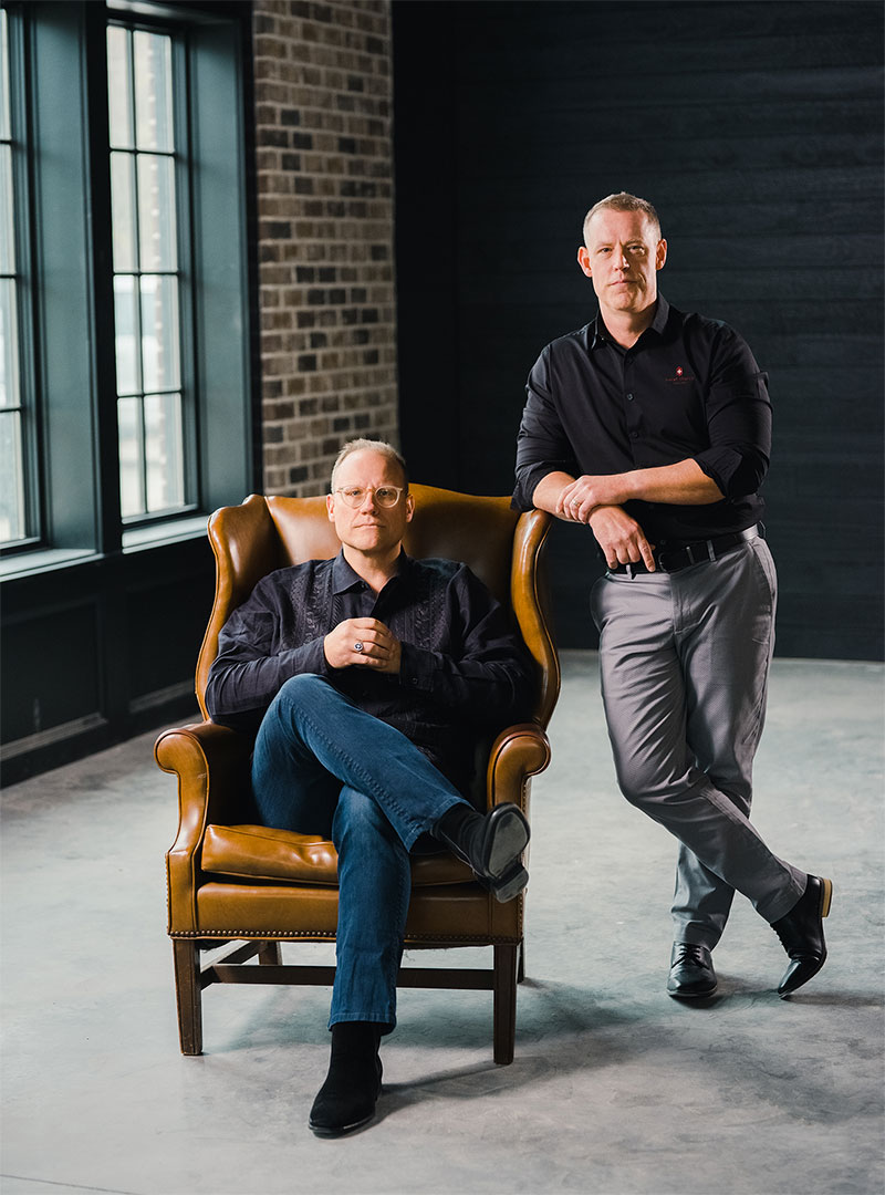 Two men posing indoors, one seated in a brown leather armchair and the other standing with arms crossed.