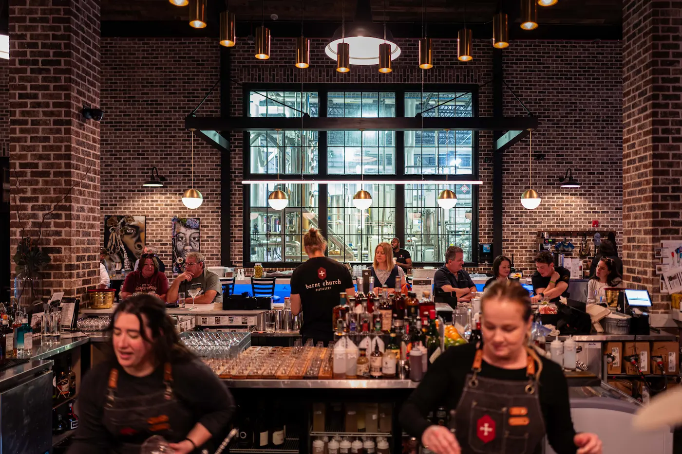 Modern distillery tasting room with brick walls, bartenders, and patrons seated at the bar.