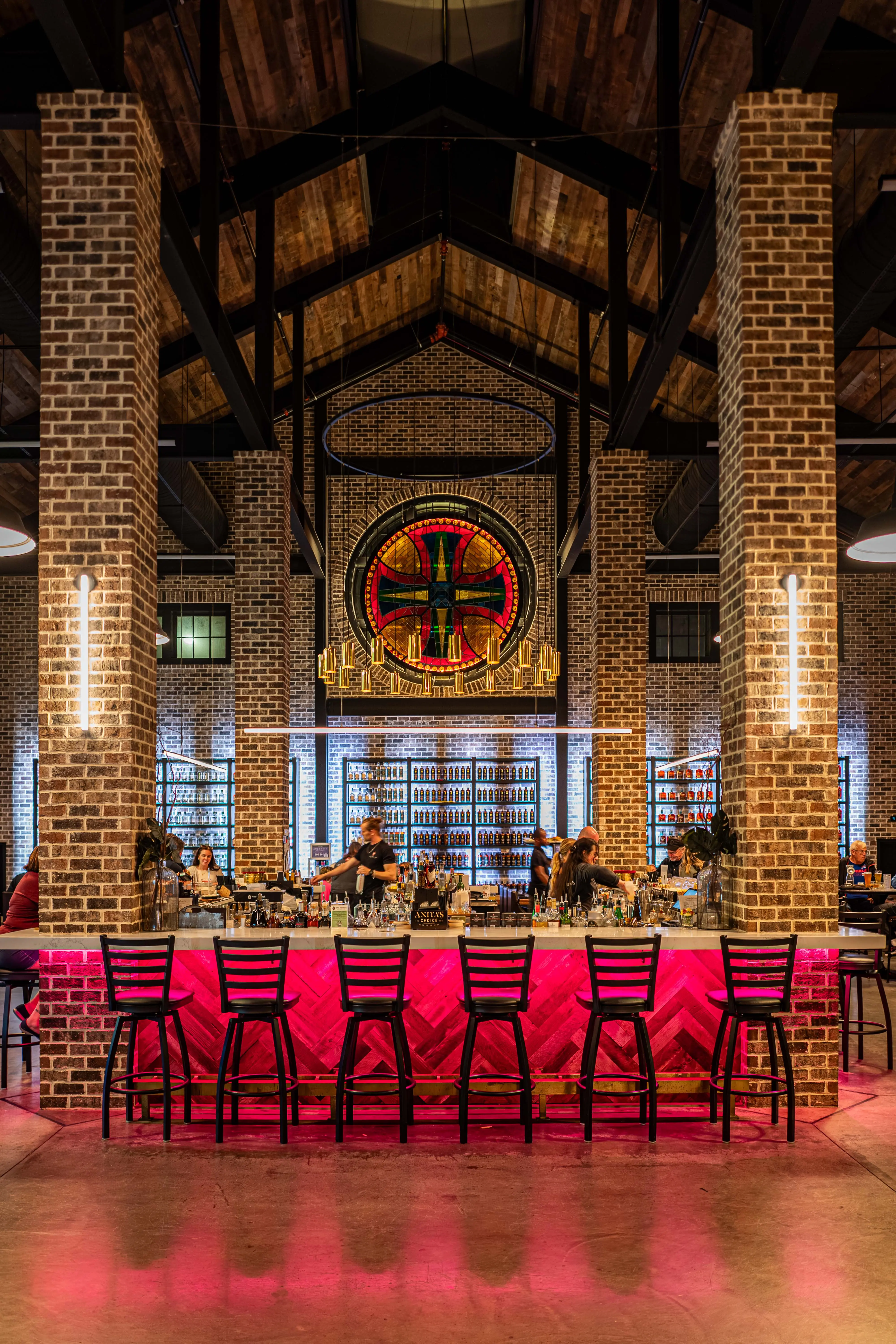 Interior of a stylish bar with exposed brick pillars, wooden ceiling, black chairs, and a lit pink bar counter.