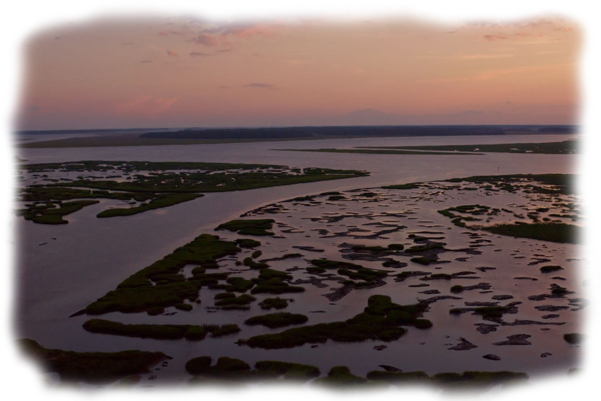 Aerial view of tidal marshlands with patches of green vegetation surrounded by water under a colorful sunset sky.