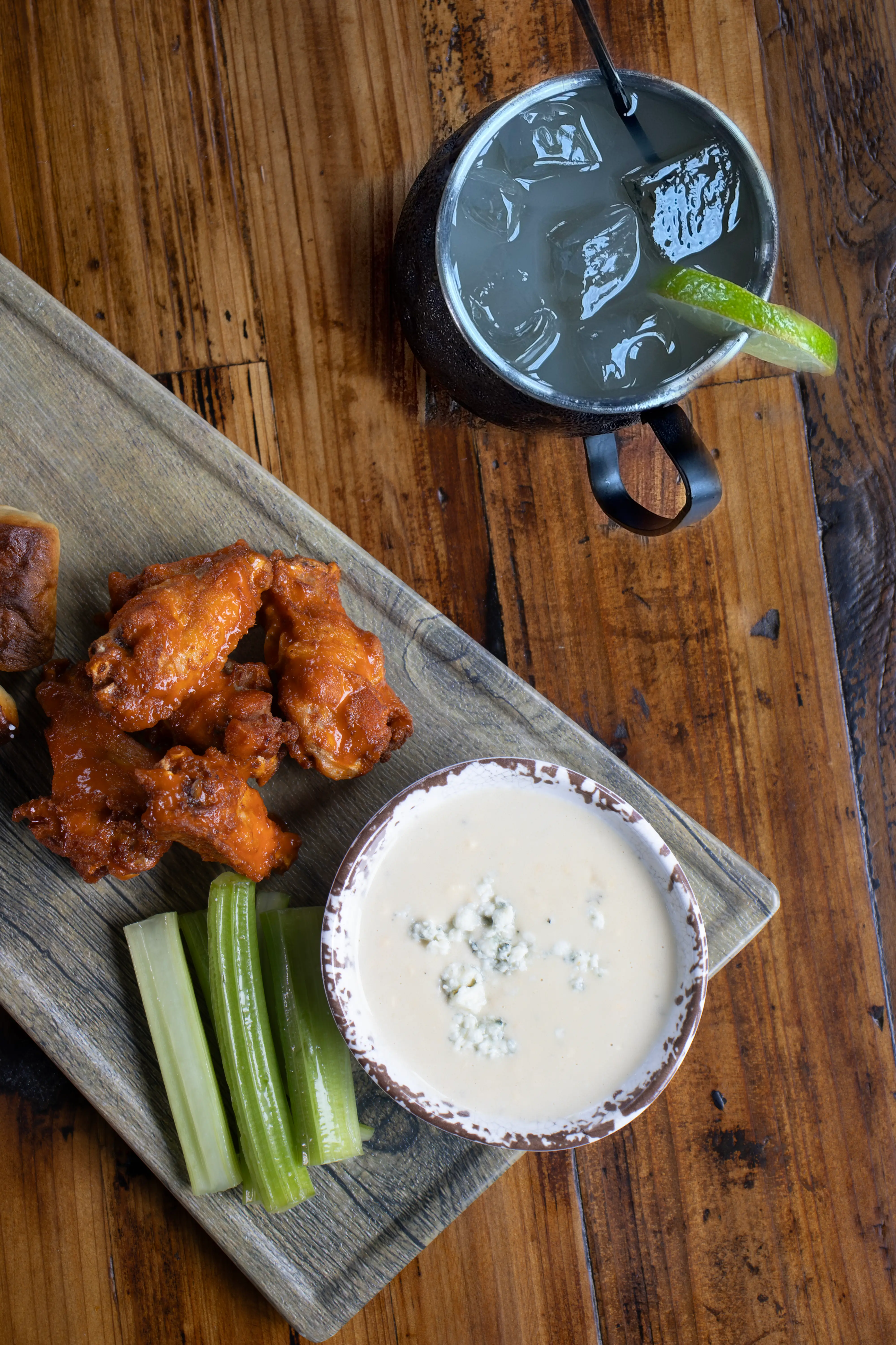 Wooden board with buffalo chicken wings, celery sticks, blue cheese dip, and a metal mug with ice and lime wedge on a wooden table.