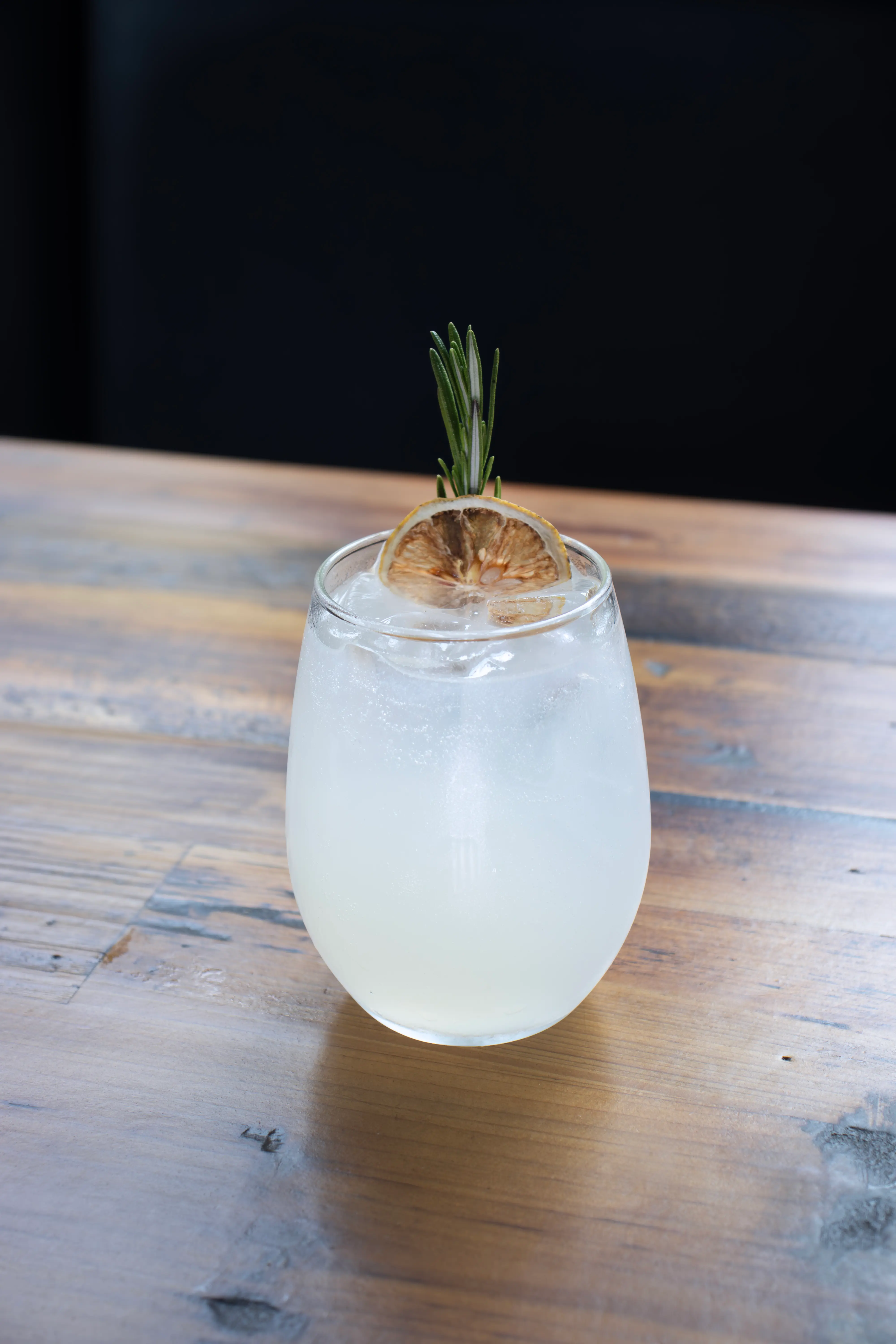 Clear cocktail with ice, garnished with a dried lemon slice and a sprig of rosemary on a wooden table.