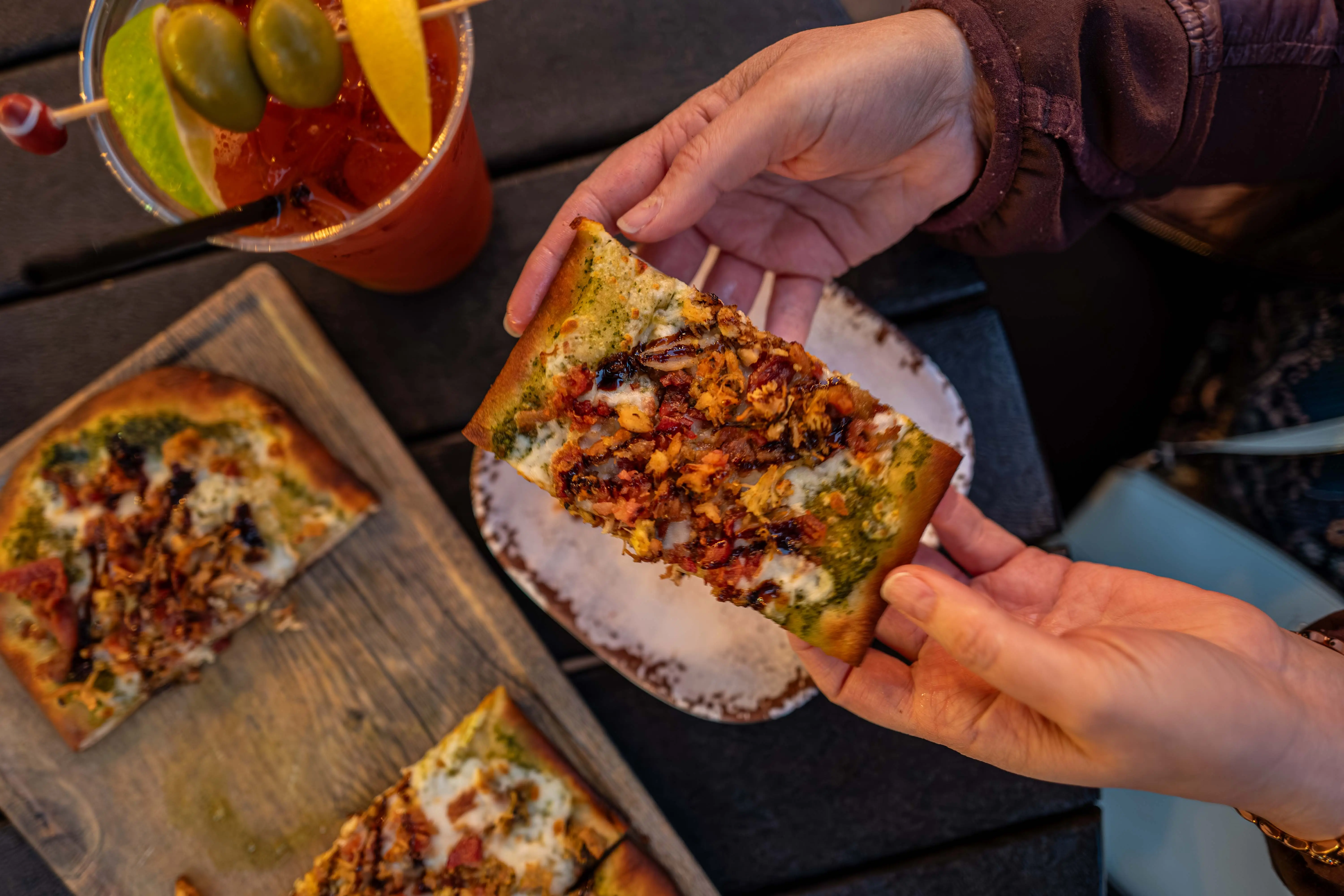 Hands holding a rectangular slice of flatbread pizza with pesto, cheese, and meat toppings, next to a Bloody Mary cocktail garnished with olives and citrus.