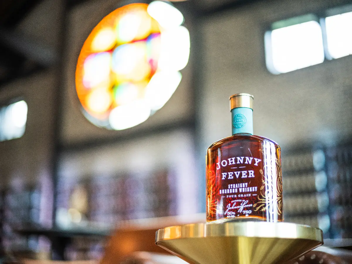 Bottle of Johnny Fever Straight Bourbon Whiskey on a brass stand in a dimly lit room with stained glass windows.