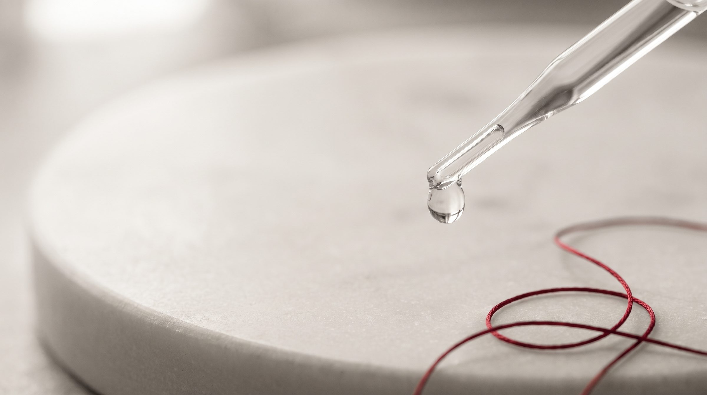 A single clear drop at the tip of a glass pipette above a pale stone surface.