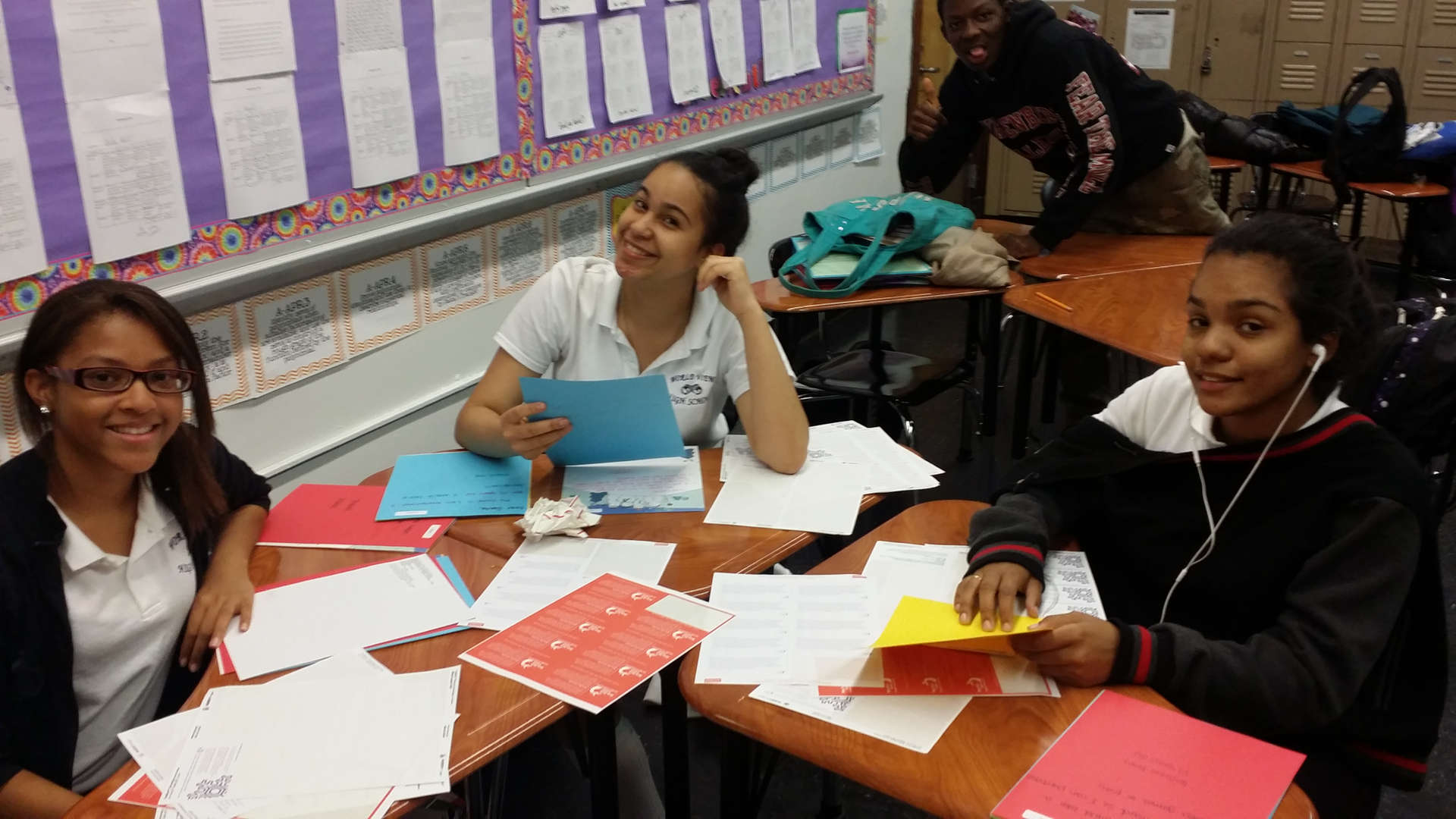 Students smiling at classroom desks covered with papers and folders