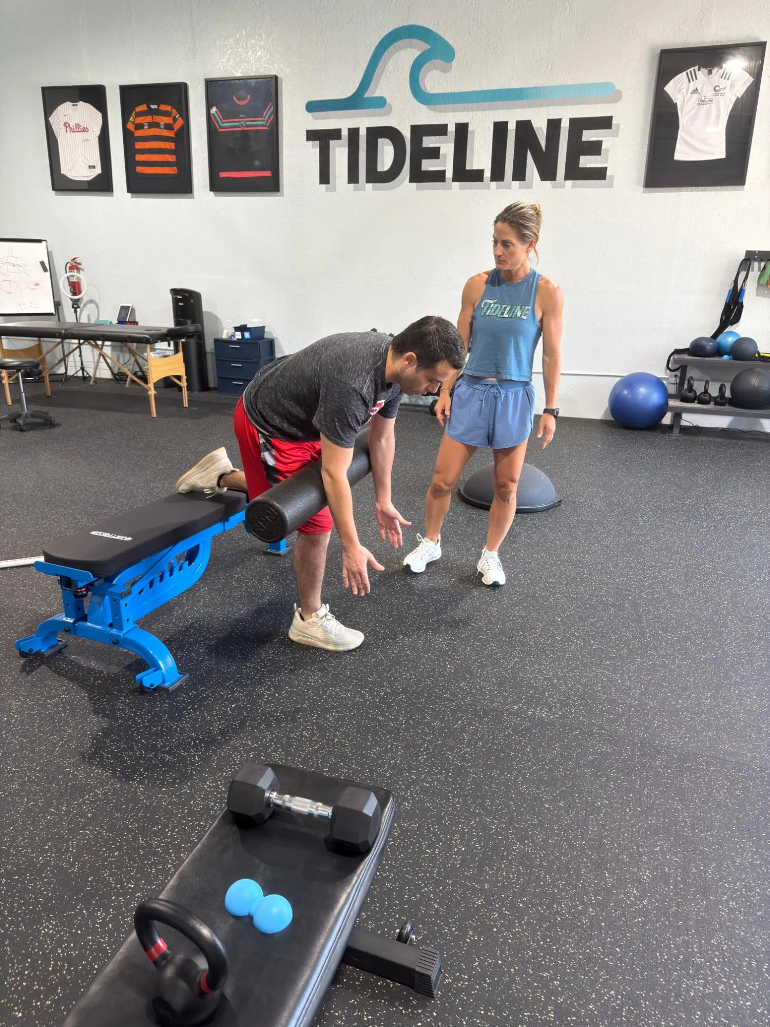 Trainer watches man using foam roller on leg supported by bench in a gym with Tideline logo.