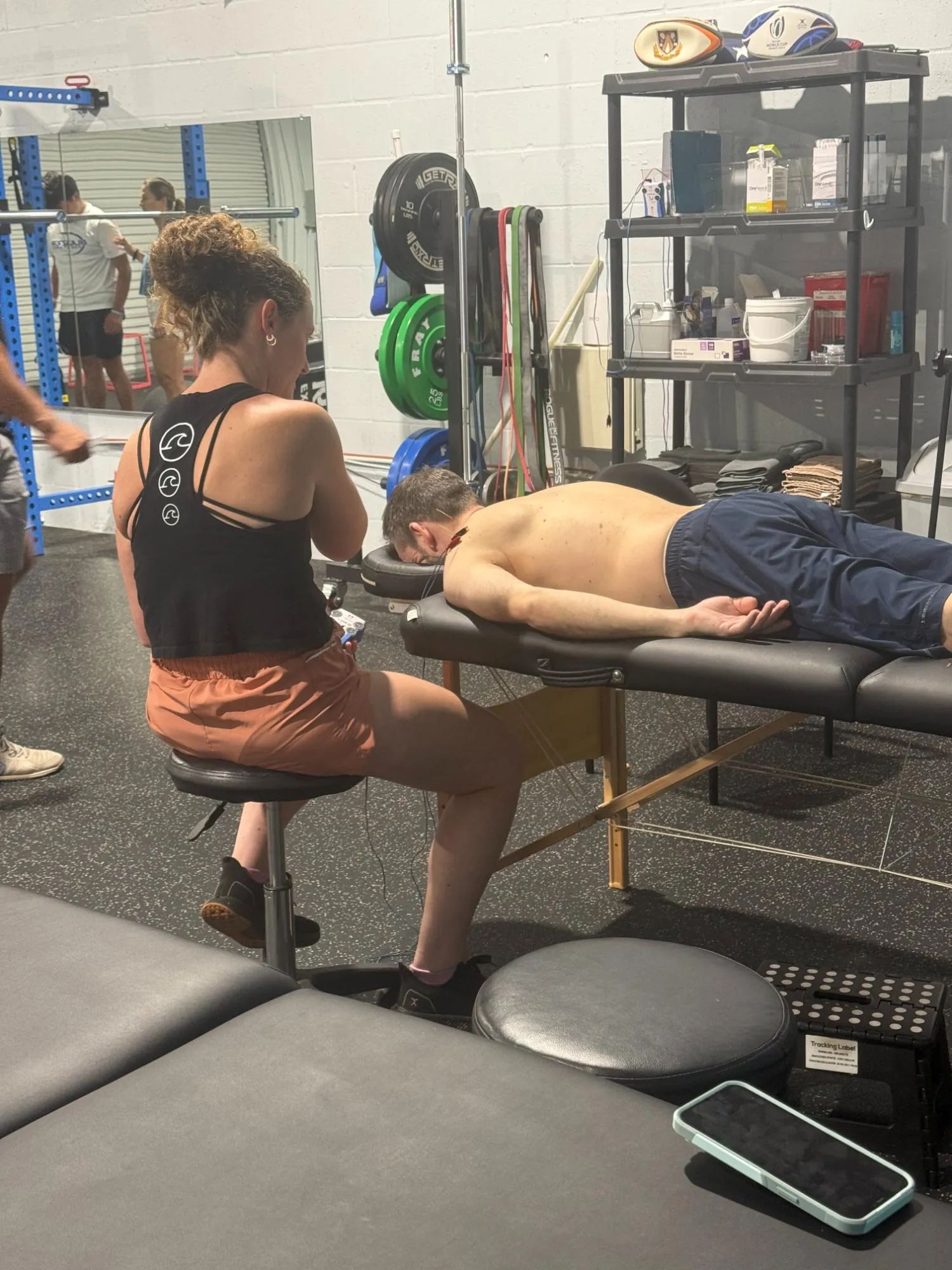Woman sitting on a stool next to a shirtless man lying face down on a massage table in a gym.