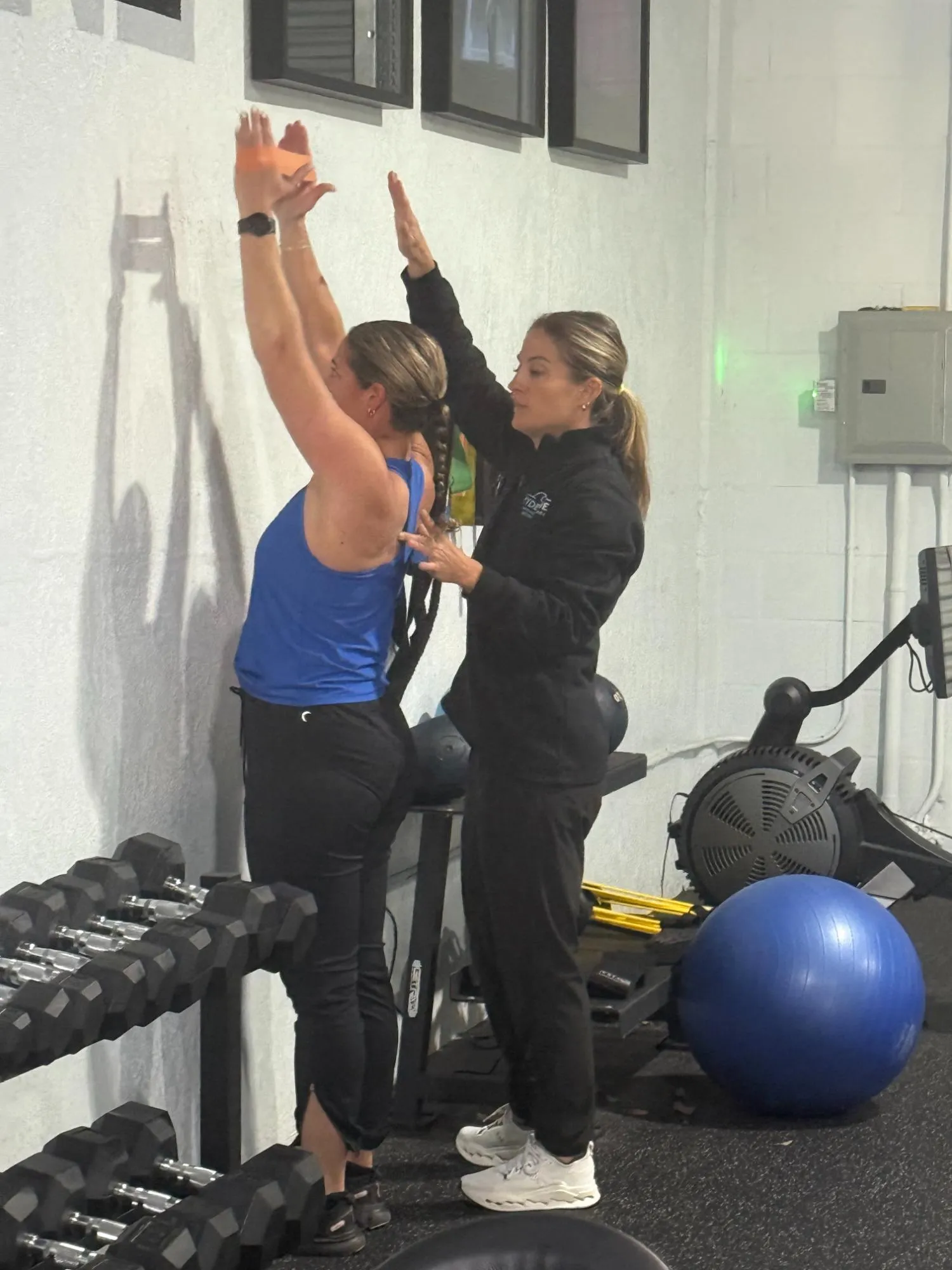 Trainer assists woman doing wall slides exercise near dumbbell rack in a gym.