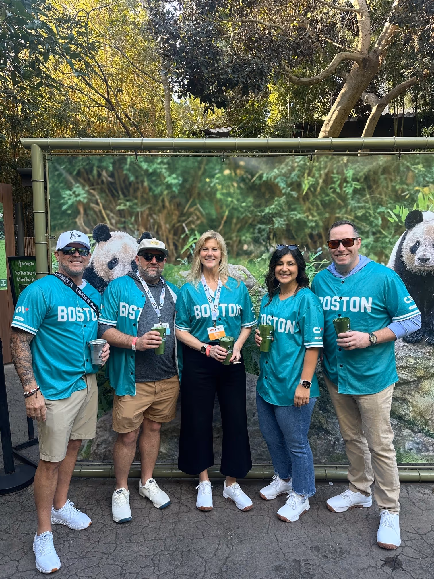 Five people in teal Boston shirts holding drinks pose in front of a panda backdrop outdoors.