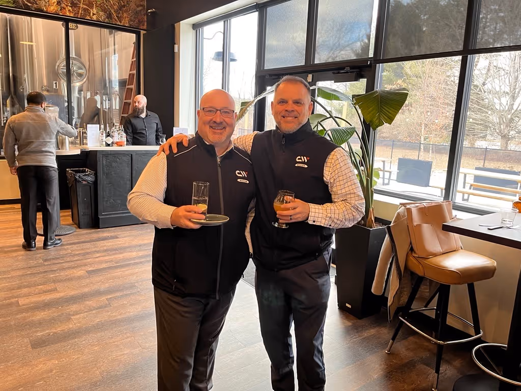 Two men wearing matching black vests smiling and holding drinks in a modern bar with large windows.
