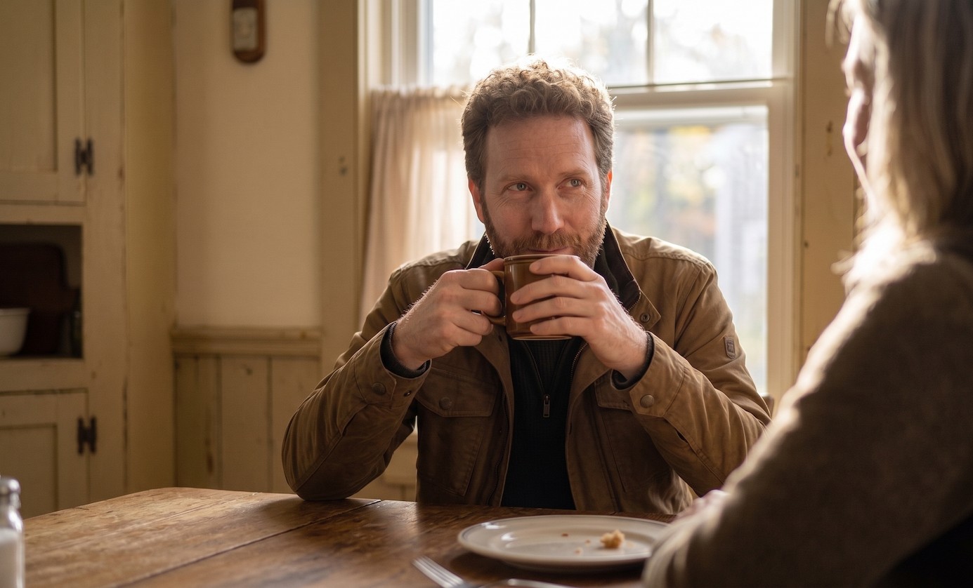 A man at a kitchen table in morning light, listening intently to someone across from him.