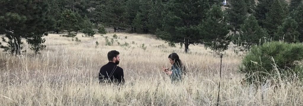 Two people sitting in tall meadow grass in conversation, representing the admissions conversation you have when choosing a coaching certification program.