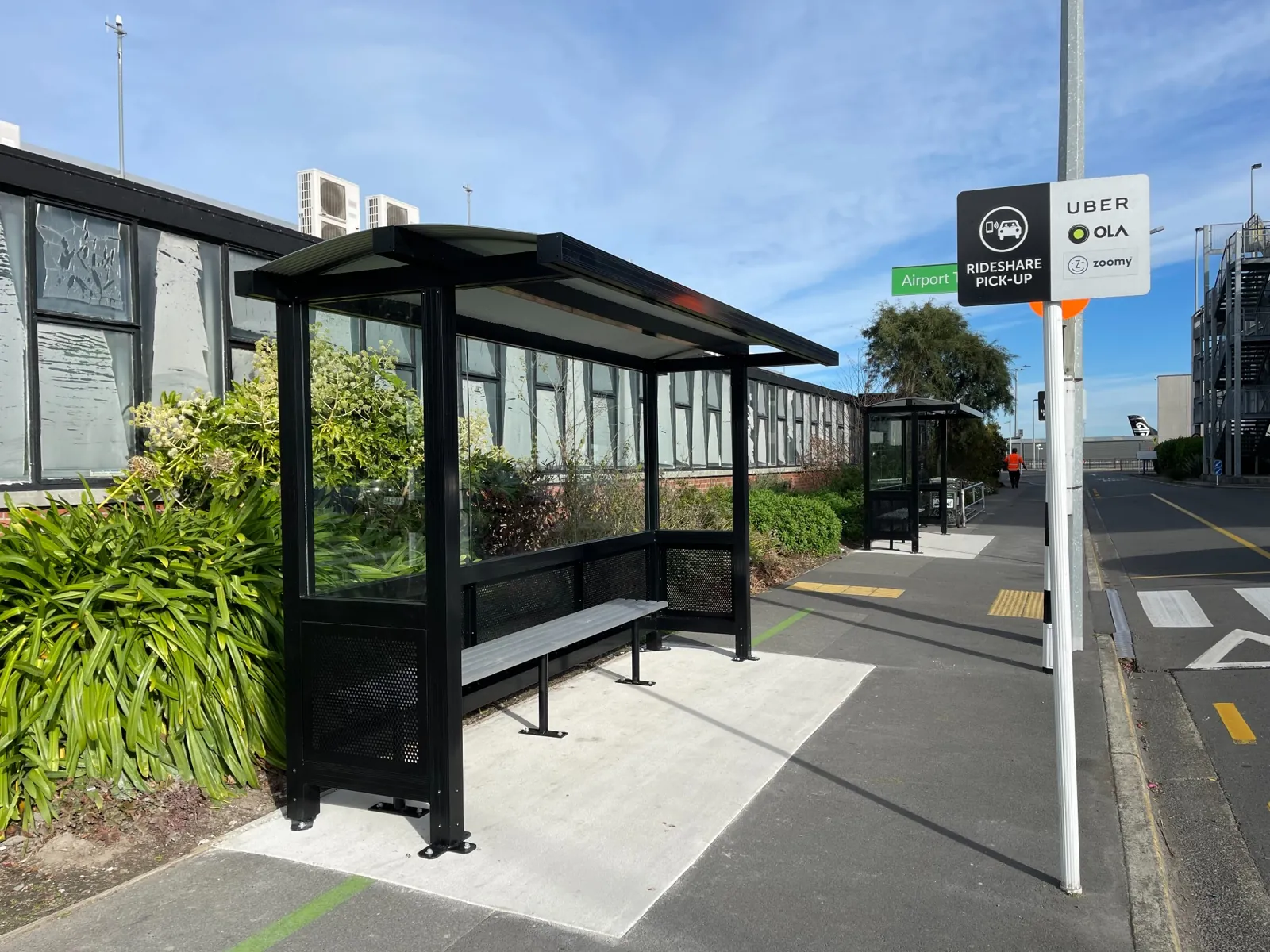 Black aluminium shelter with glass and perforated panels beside an airport pick-up area