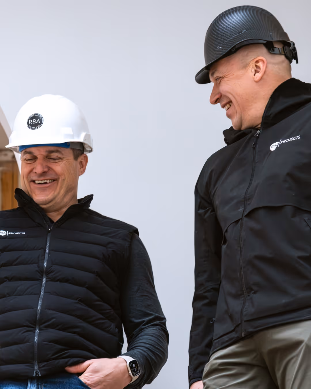 Two men on a construction site smiling and wearing safety helmets and black jackets branded with 'RBA Projects'.