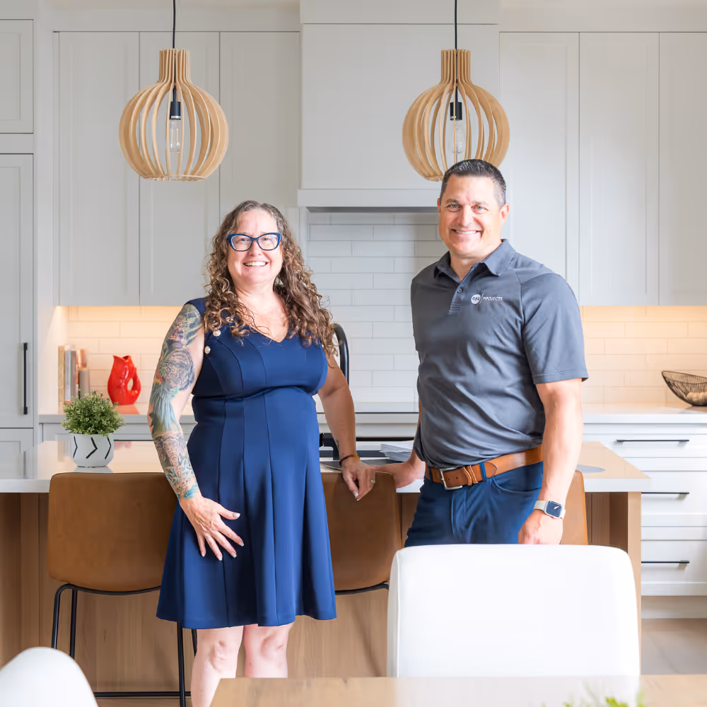 Two people standing and smiling in a modern kitchen with white cabinets, wooden pendant lights, and a kitchen island with brown chairs.