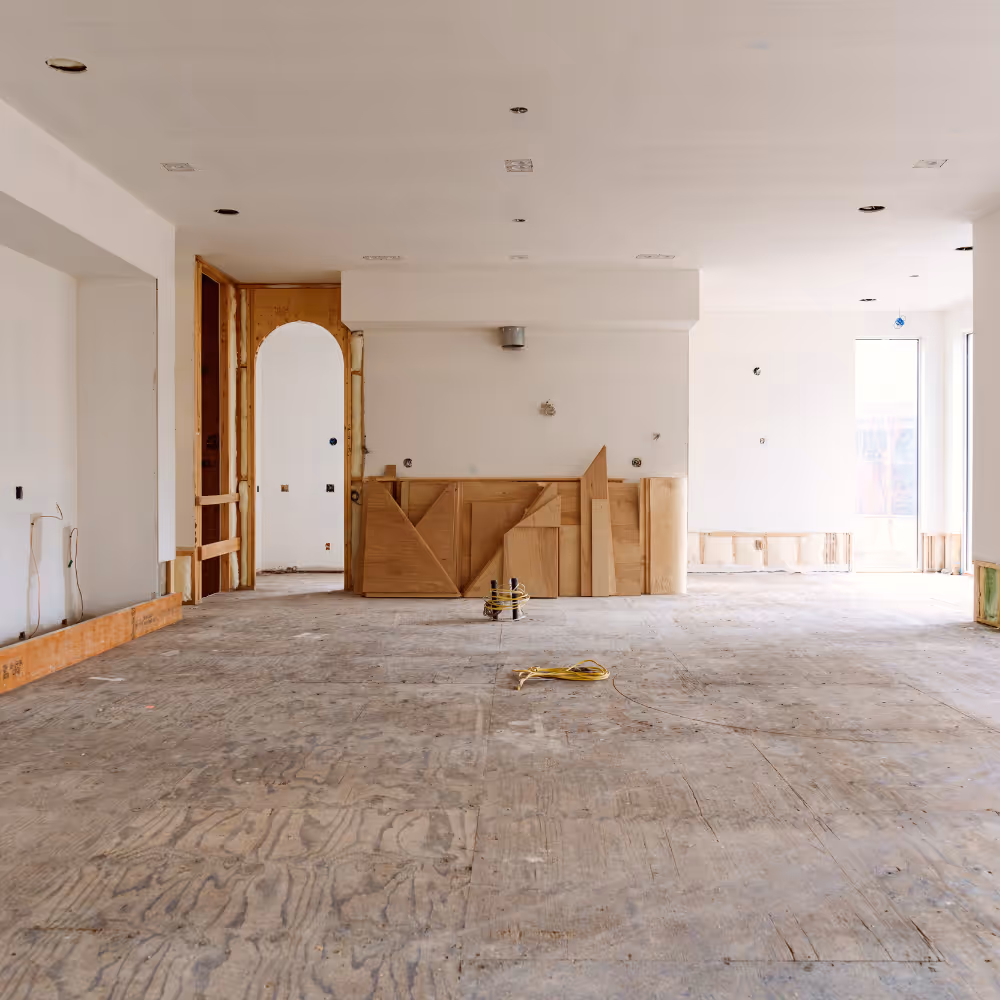 Large empty room under renovation with bare plywood floor, white walls, unfinished wooden framing around doorways, and some construction materials on floor.