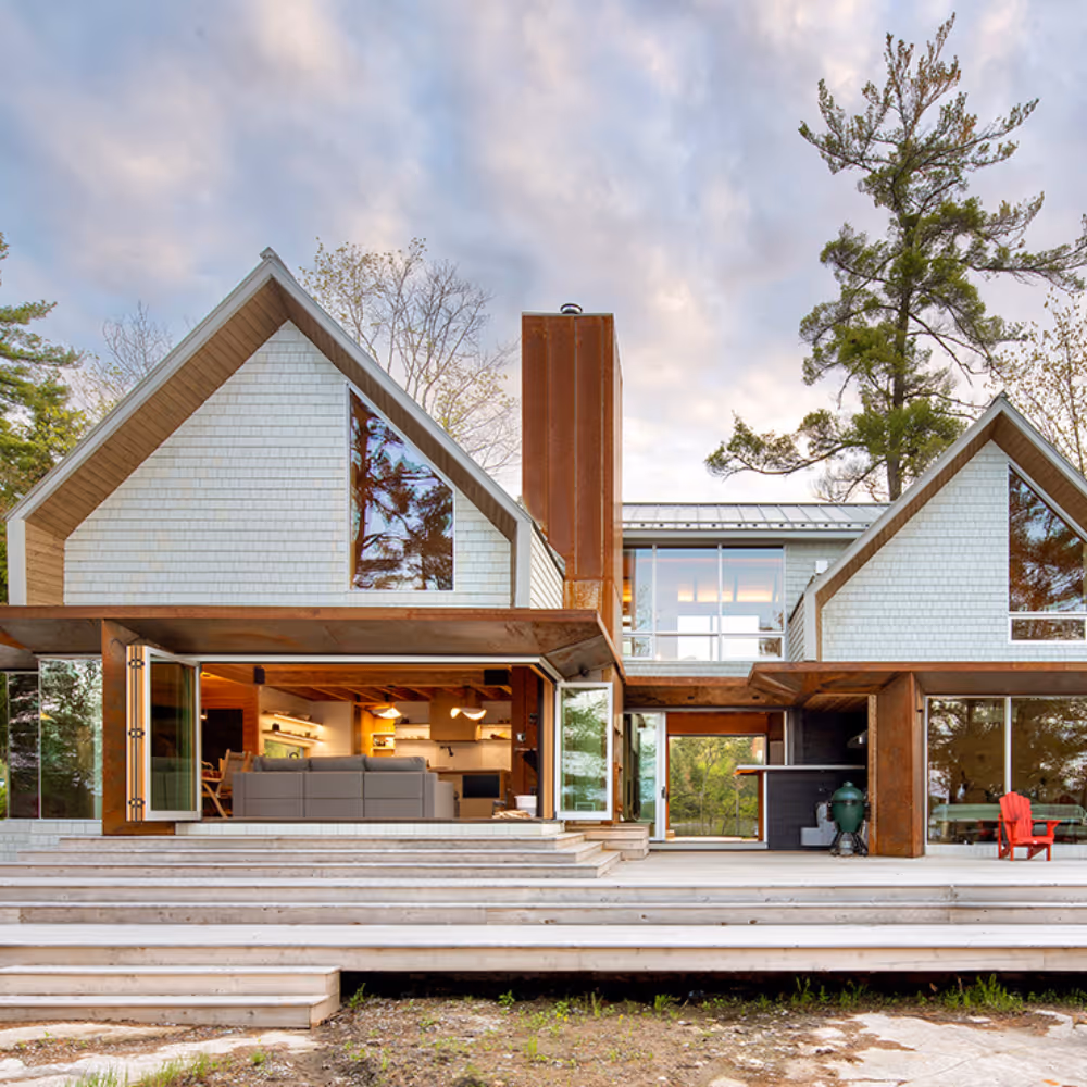 Modern two-story house with large triangular windows, white siding, and rust-colored metal accents, featuring open glass doors revealing interior living space, wide wooden steps leading to a deck, surrounded by trees under a cloudy sky.