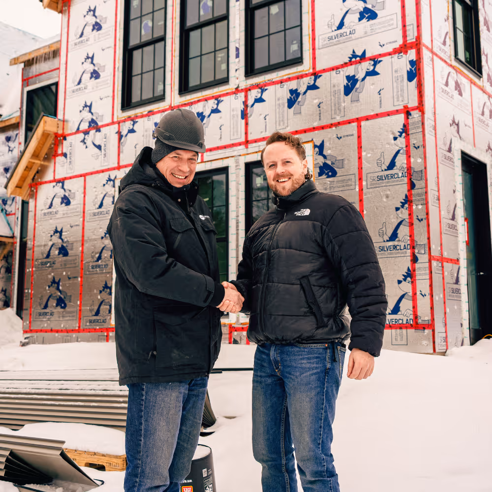 Two men shaking hands and smiling in front of a partially constructed house wrapped with Silverclad insulation panels in a snowy setting.