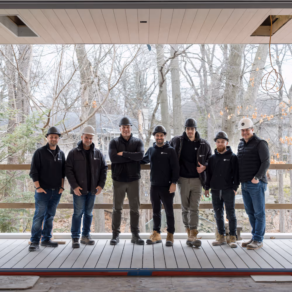 Seven construction workers standing side by side on a wooden deck or platform, wearing work boots and helmets, with leafless trees and houses visible outside through a large open window or frame behind them.
