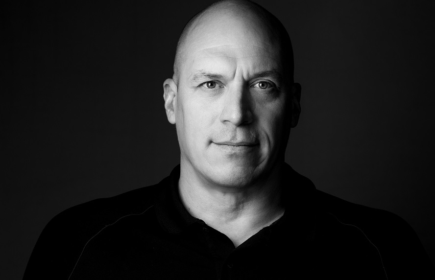 Black and white portrait of a middle-aged bald man with light eyes, wearing a dark collared shirt, looking directly at the camera against a dark background.