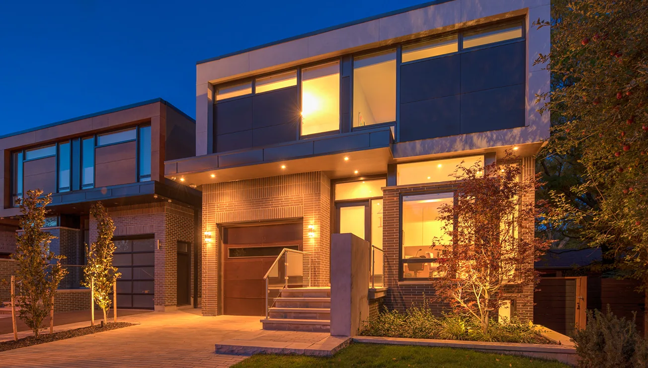 Modern two-story house at dusk with large windows, brick and panel exterior, a front porch with stairs and glass railing, illuminated by warm interior and exterior lights, surrounded by small trees and landscaping.