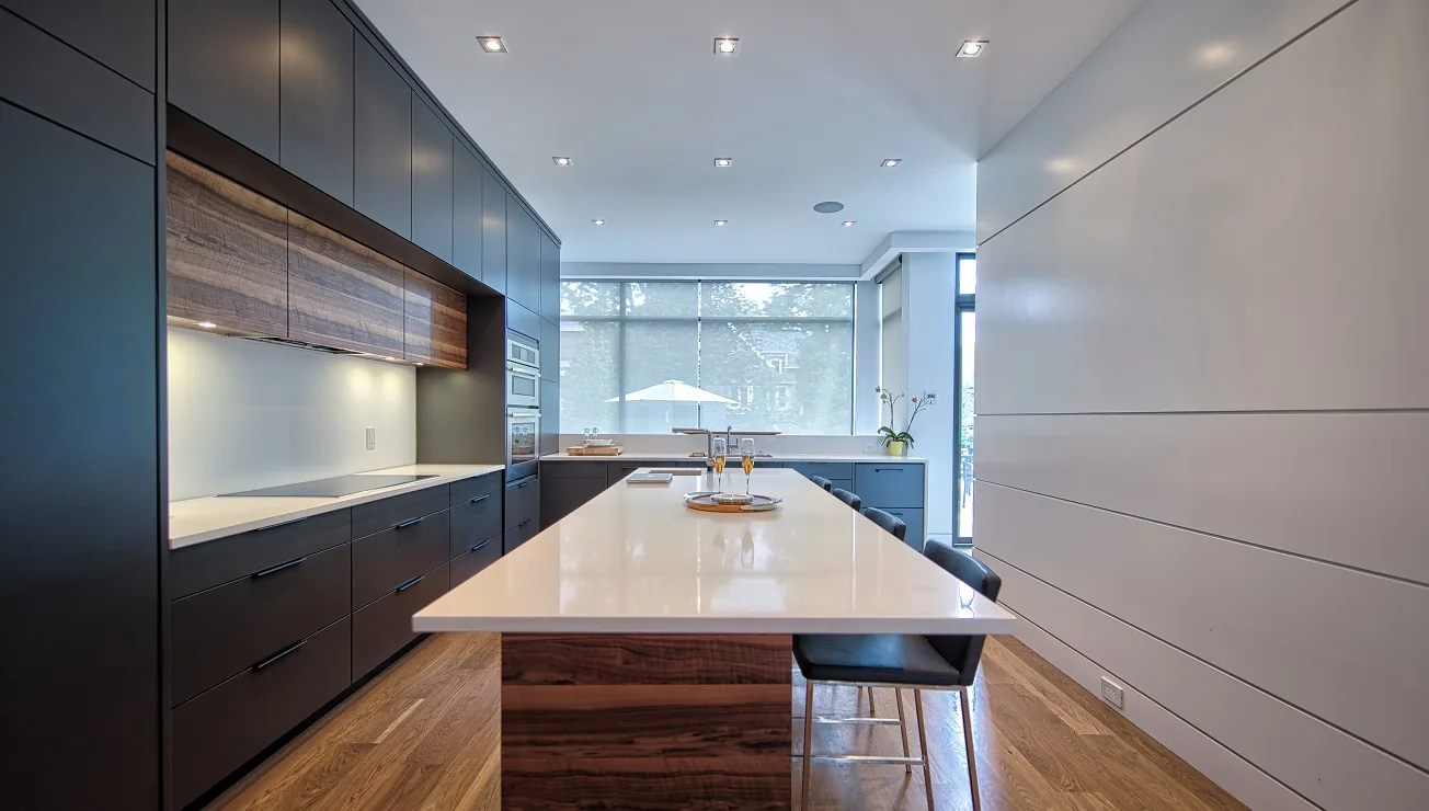 Modern kitchen with dark cabinetry, a large white island countertop with bar stools, wooden flooring, and a window with a blind letting in natural light.